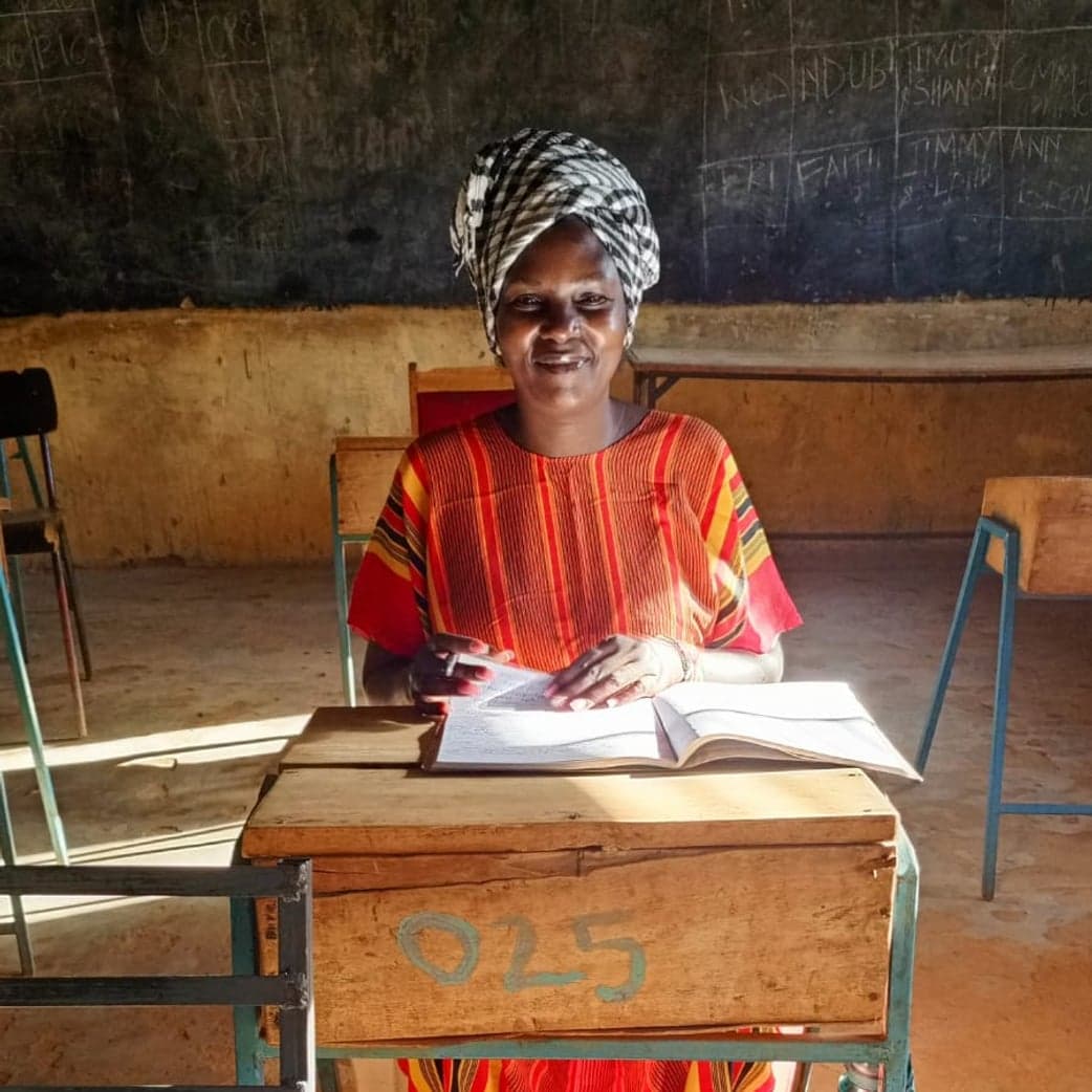 A smiling woman in a headscarf seated at a classroom desk with the group record book open