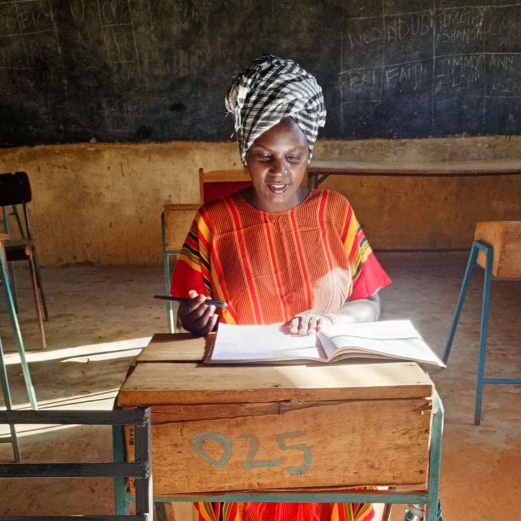 A woman in a headscarf, pen in hand, reading the group record book at a classroom desk